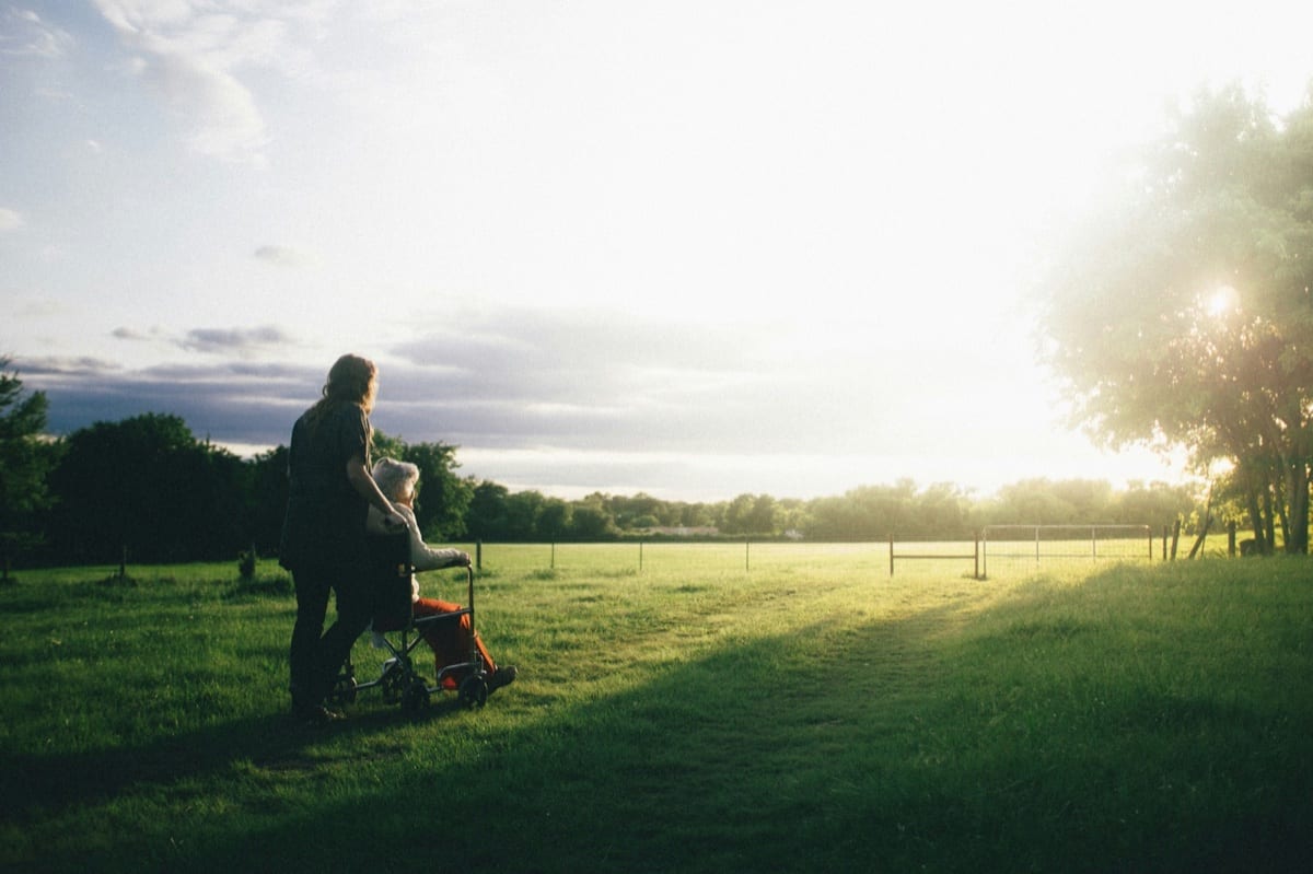 A caregiver pushing someone in a wheelchair across a grassy field at sunset